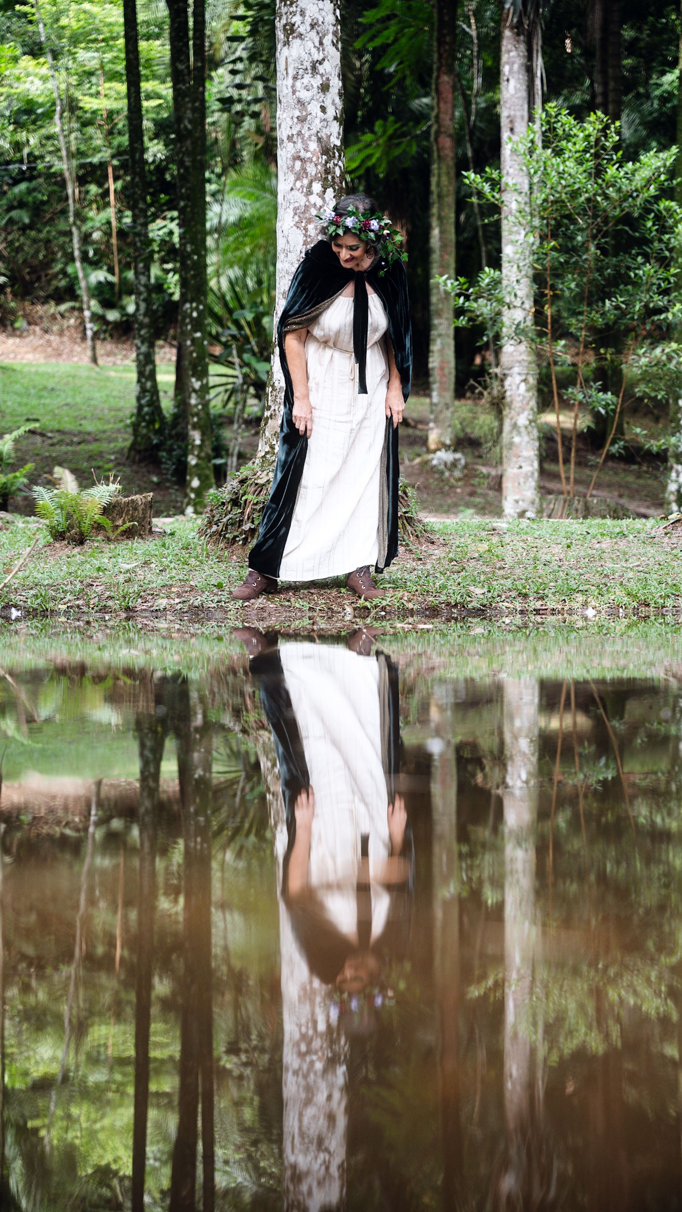 A woman in a forest reflects in a pond, wearing a floral crown and cape.