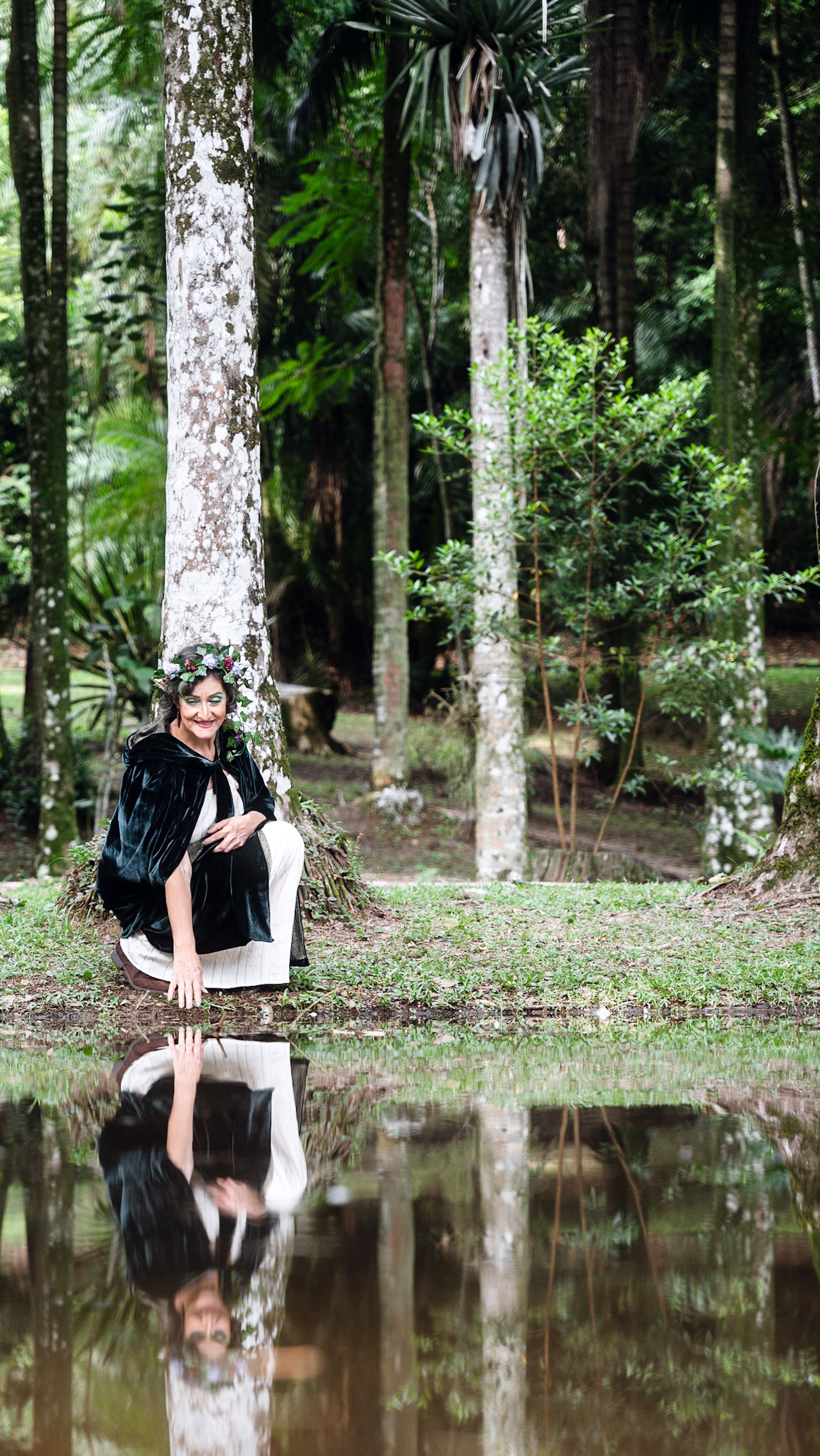 A woman sits by a forest pond with her reflection visible in the water.
