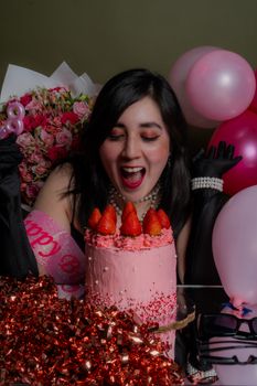 A woman joyfully celebrating her birthday with a pink strawberry cake and balloons.