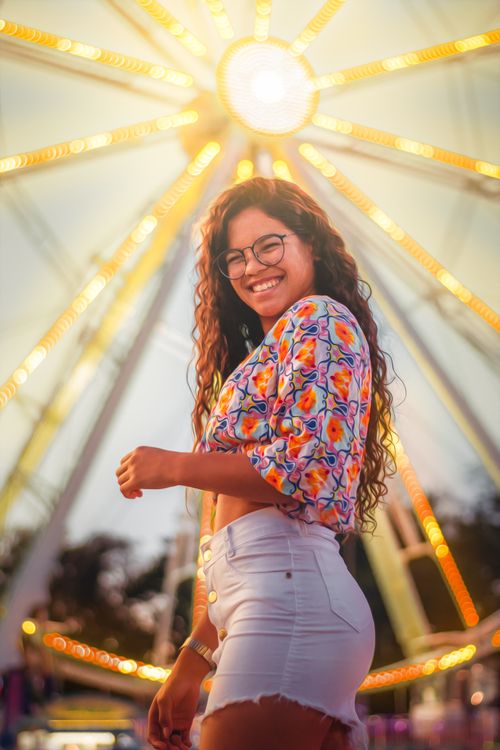 Free Happy woman with curly hair and glasses posing by a Ferris wheel at sunset. Stock Photo