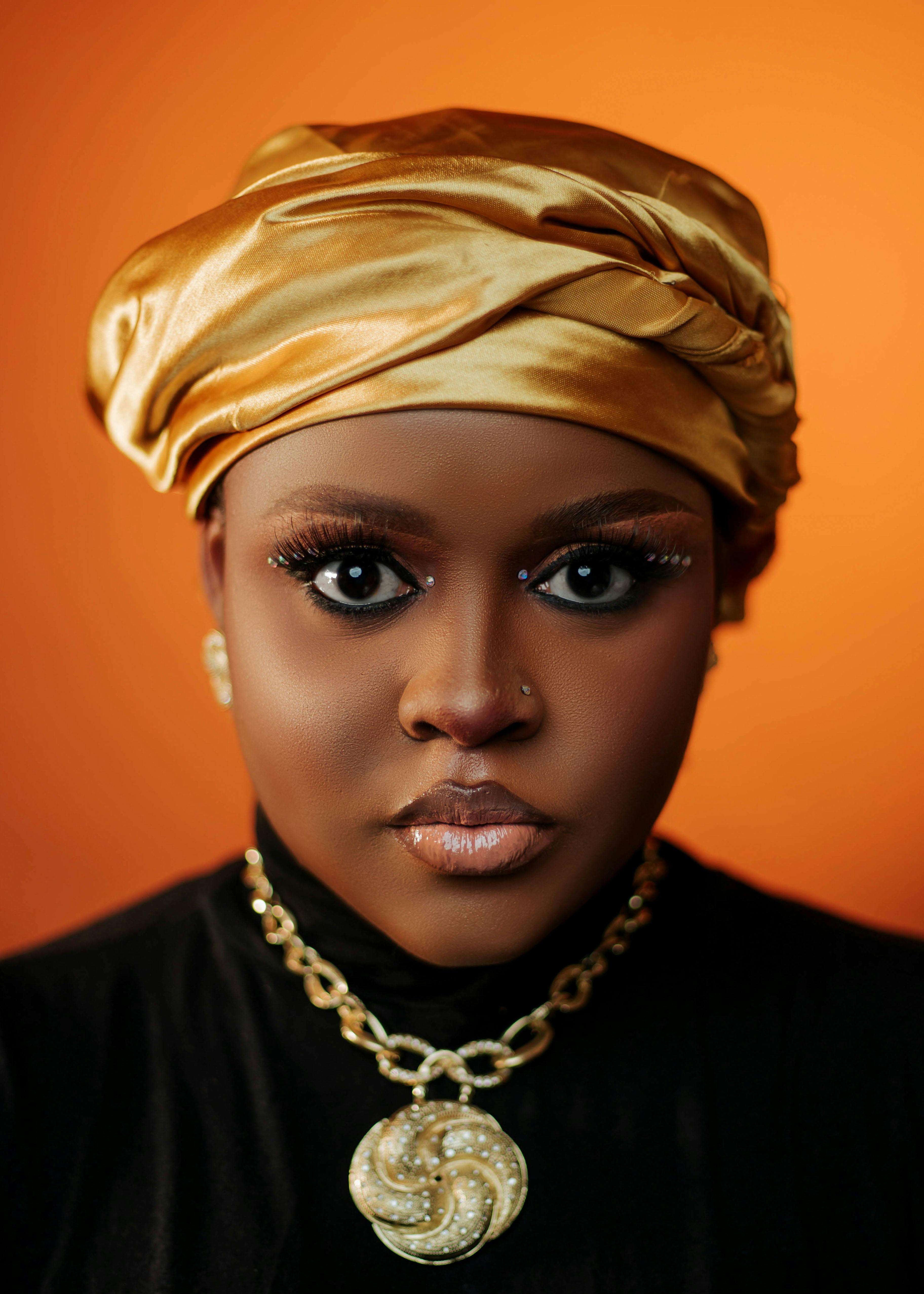 grátis Retrato em close-up de uma mulher com um lenço dourado na cabeça sobre um fundo laranja. Foto profissional
