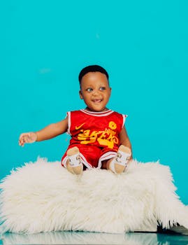 Adorable baby sitting on a fluffy white stool wearing a red basketball outfit with a vibrant turquoise background.