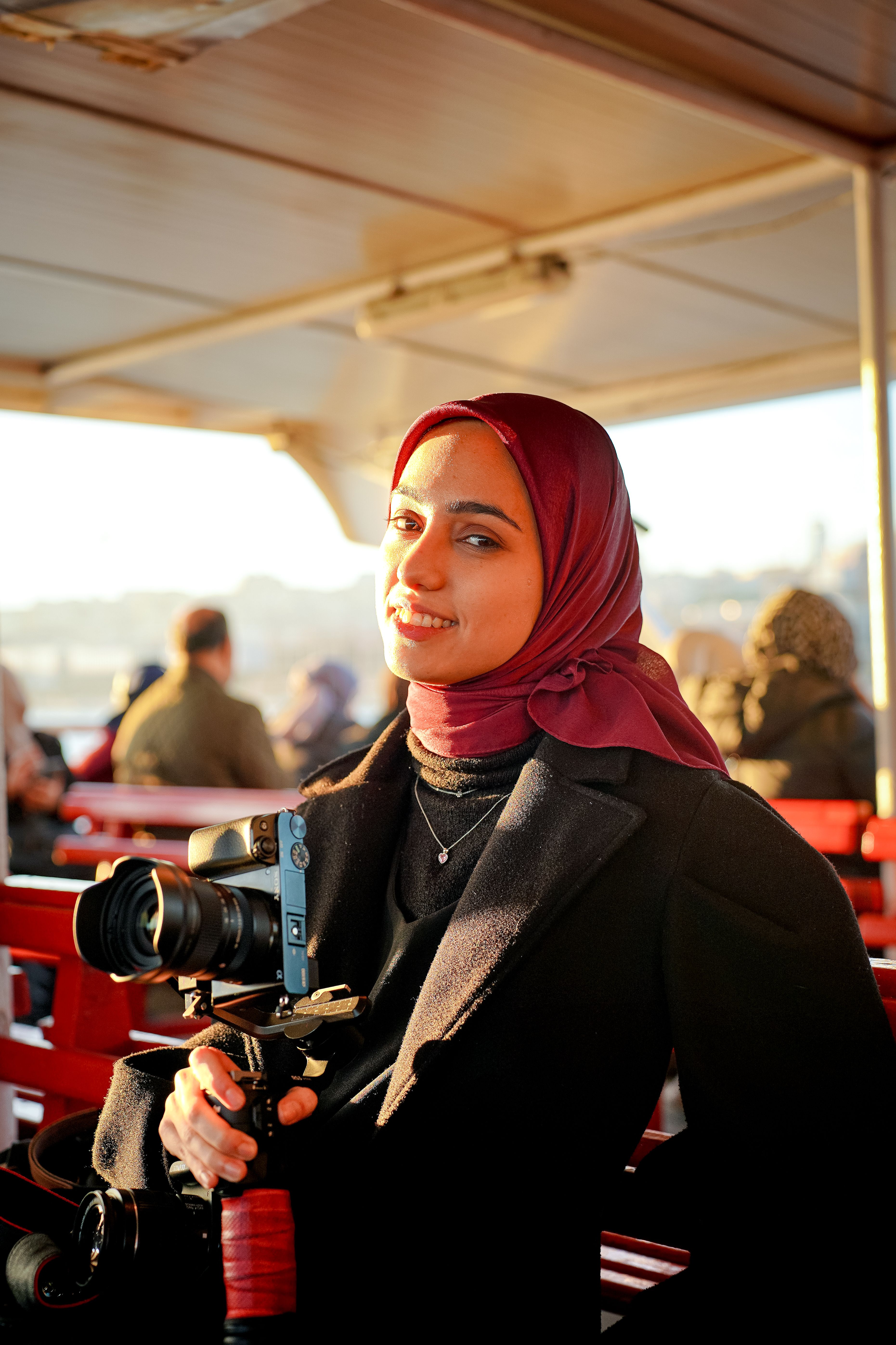 Woman in a hijab holding a camera on a boat, warmly lit setting.