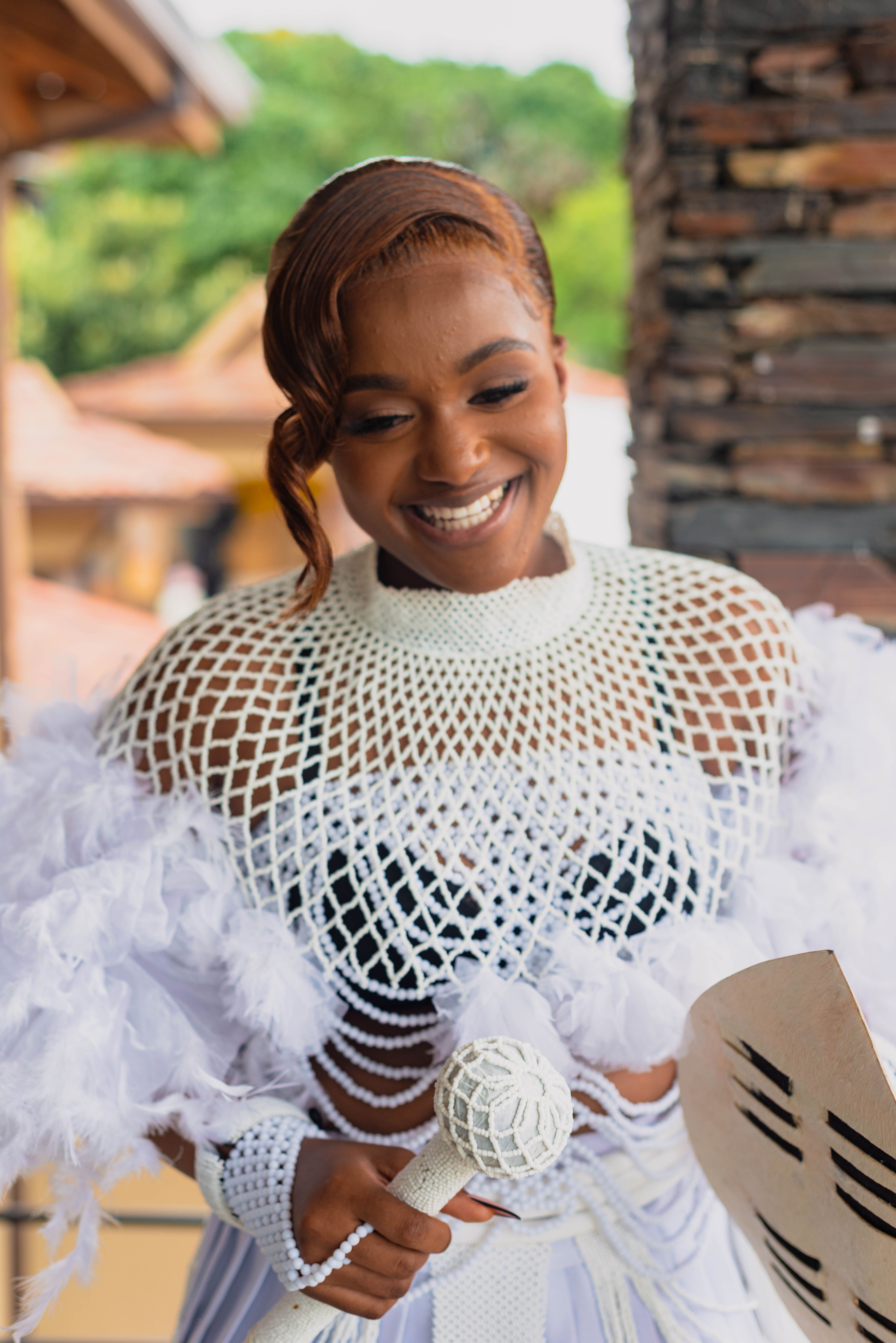 Smiling woman in a white mesh and feather costume holding a decorated scepter outdoors.