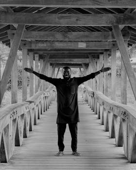 Black and white photo of a man with outstretched arms standing on a wooden bridge in an outdoor setting.