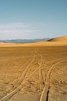 Expansive desert view featuring tire tracks, vast sand dunes, and distant mountains under a clear blue sky.