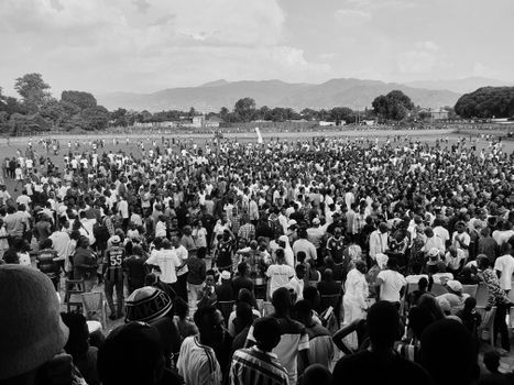 A black and white image of a large crowd gathered in an open field with mountains in the background.