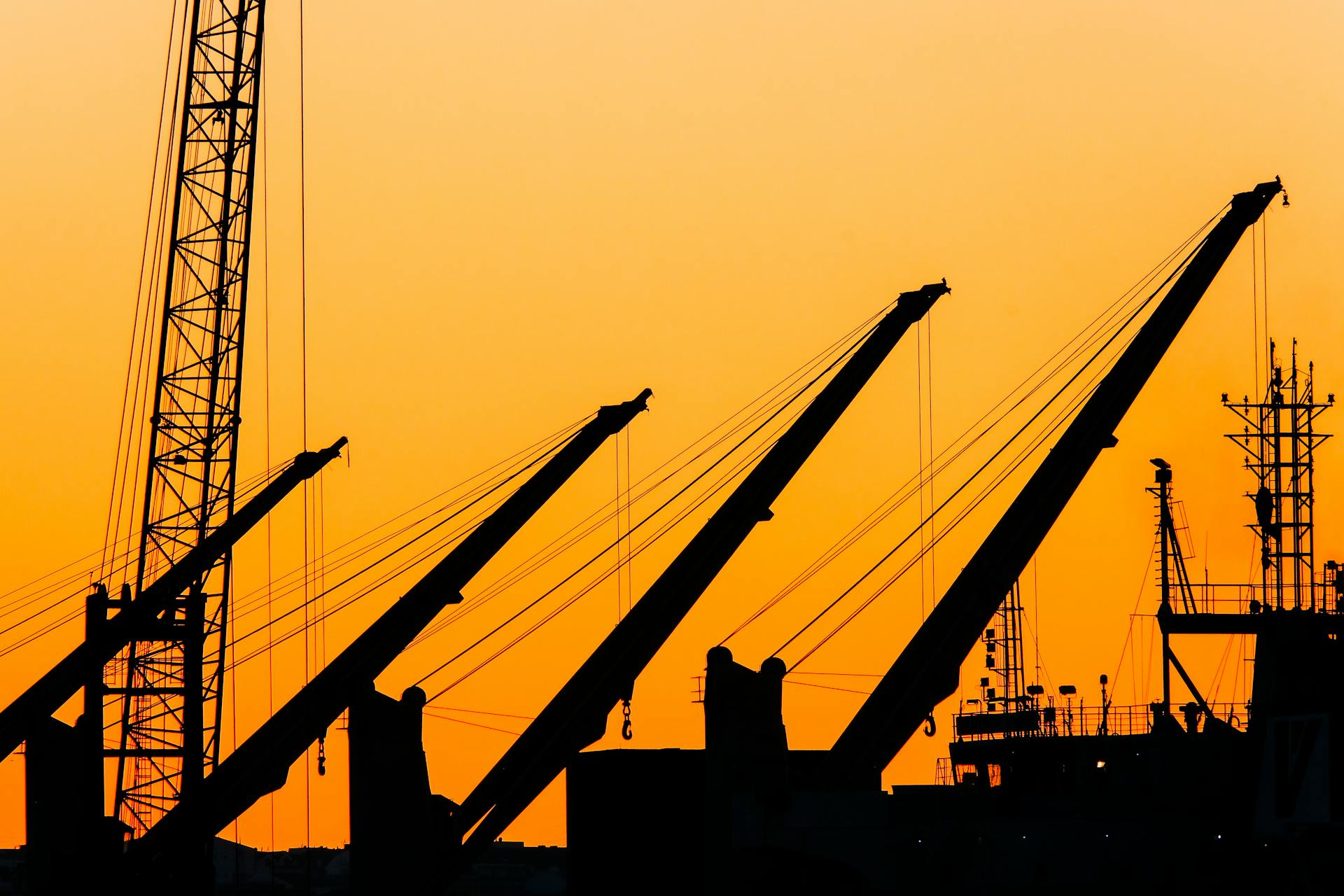 Silhouetted cranes and masts create a striking industrial skyline against a vibrant sunset backdrop.