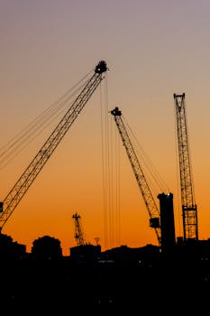 Silhouetted construction cranes against a vibrant sunset sky in an urban landscape.