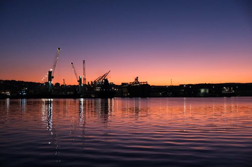 Free A stunning twilight industrial harbor scene with cranes and reflections on water under a colorful sky. Stock Photo
