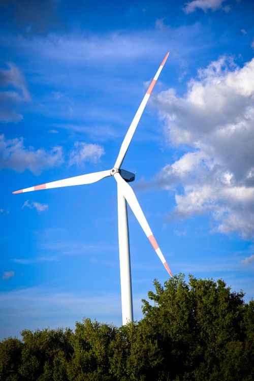 Free A wind turbine stands tall against a backdrop of a vivid blue sky with scattered clouds and green foliage. Stock Photo