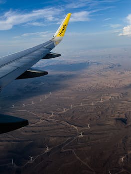 Stunning aerial view of airplane wing over desert landscape with windmills and clear blue sky.
