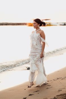 Woman in a white lace dress walking on the beach at sunset, exuding elegance.
