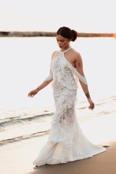A woman in a lace white dress walks along a sandy beach at sunset, embodying serenity and elegance.