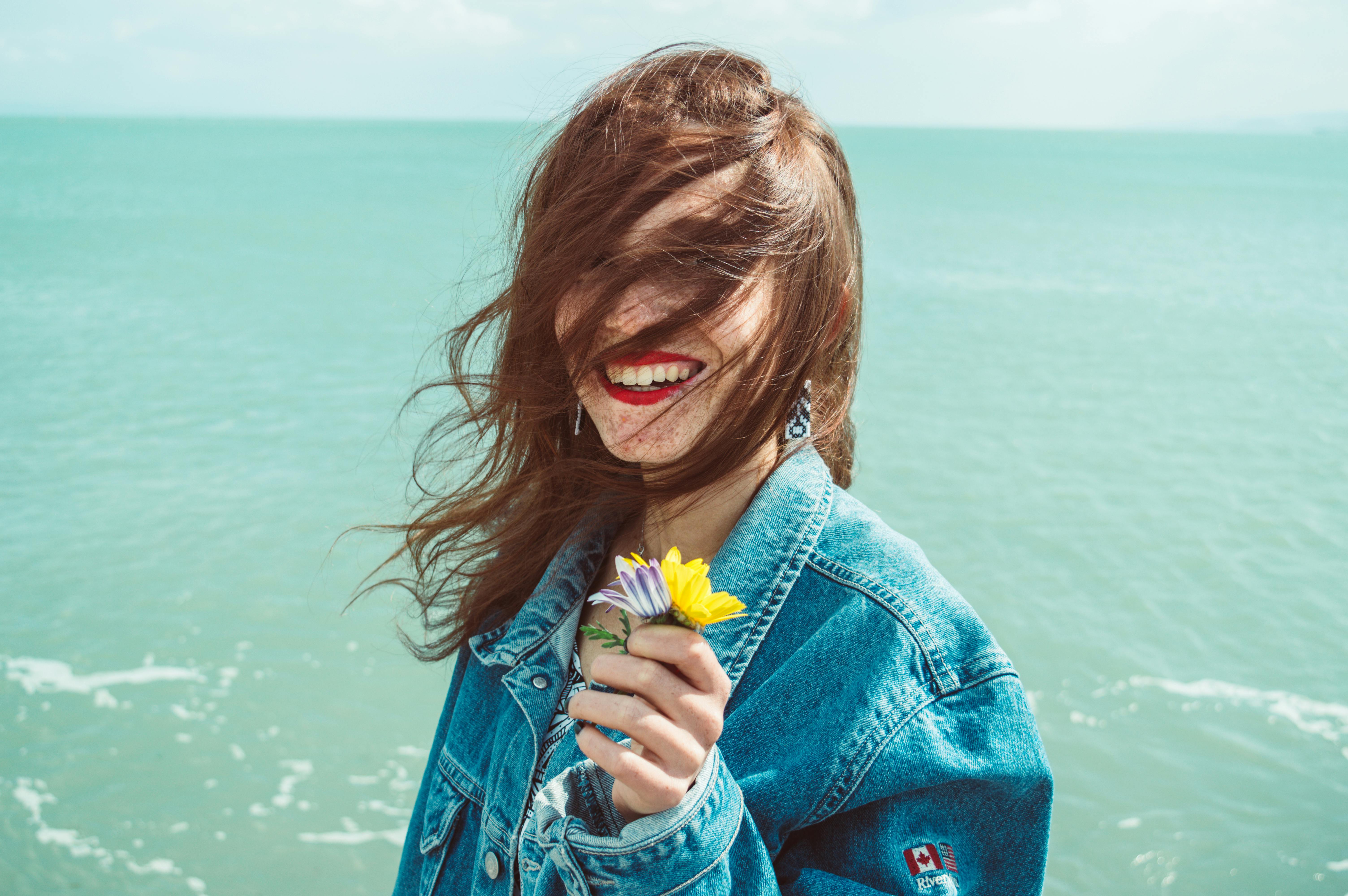 海边，一个快乐的女人穿着牛仔夹克，头发随风飘动，身旁是花朵
