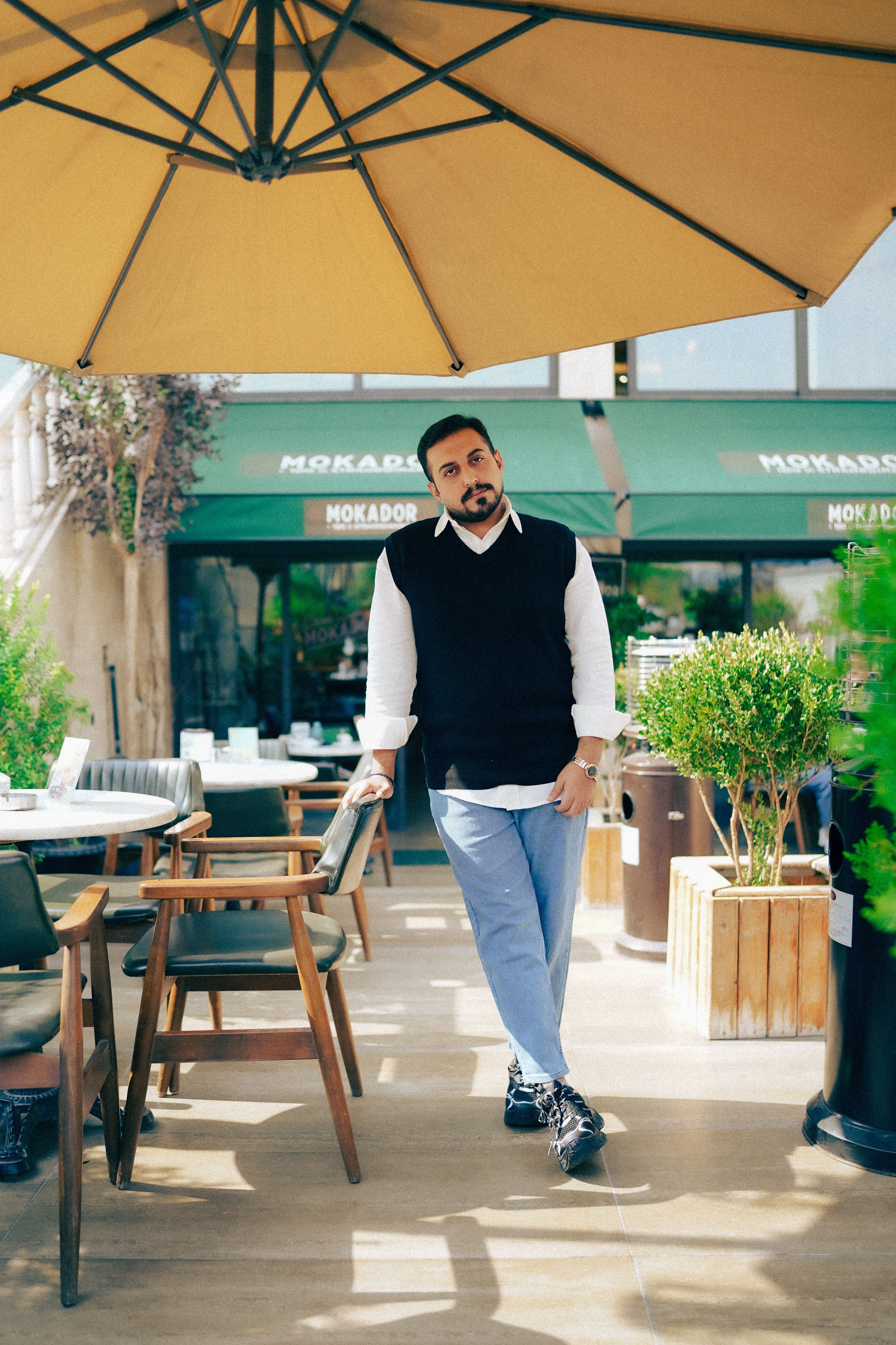 Man standing under umbrella at outdoor cafe, casual attire.
