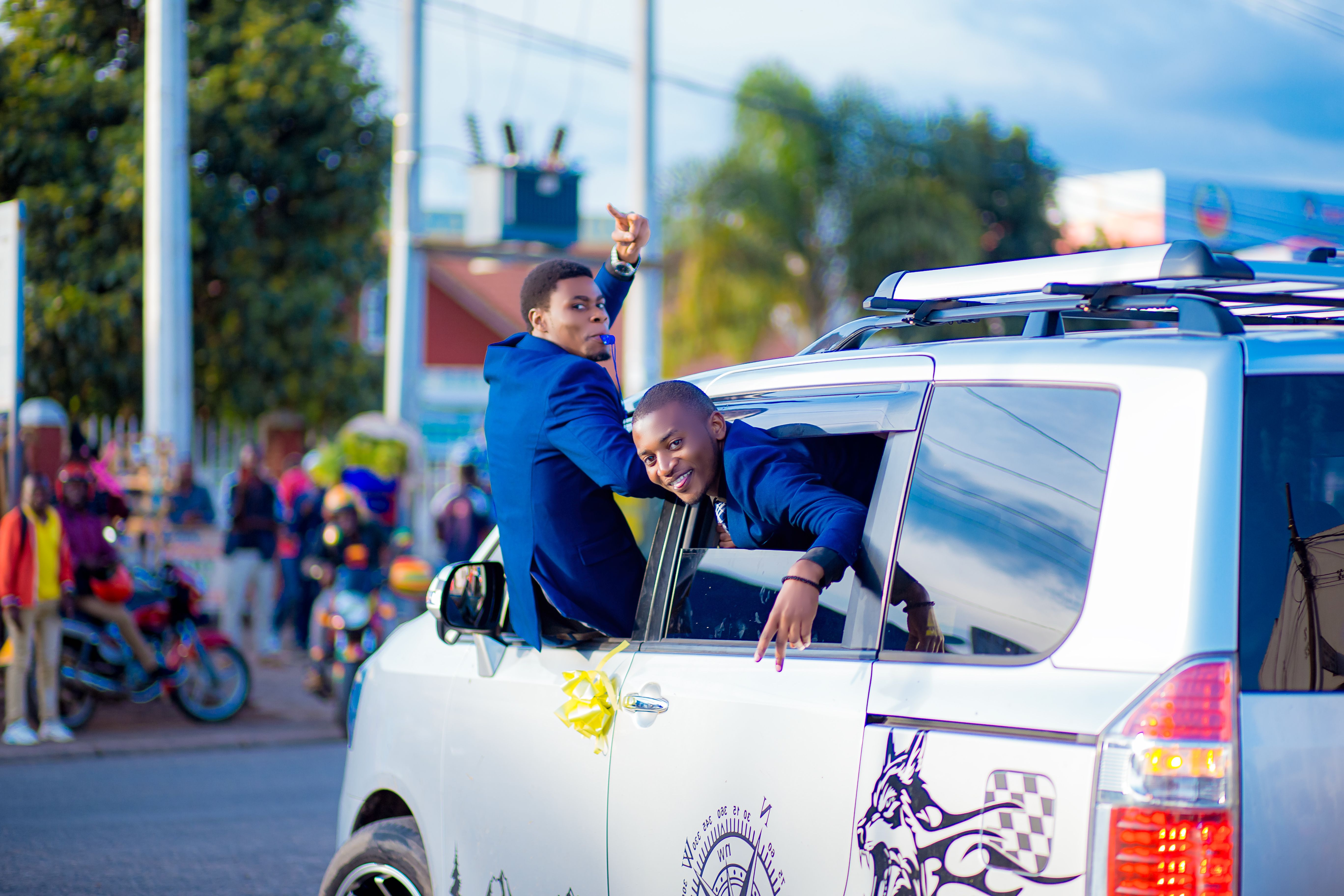 Two young men celebrating from a moving car window in a vibrant urban scene.