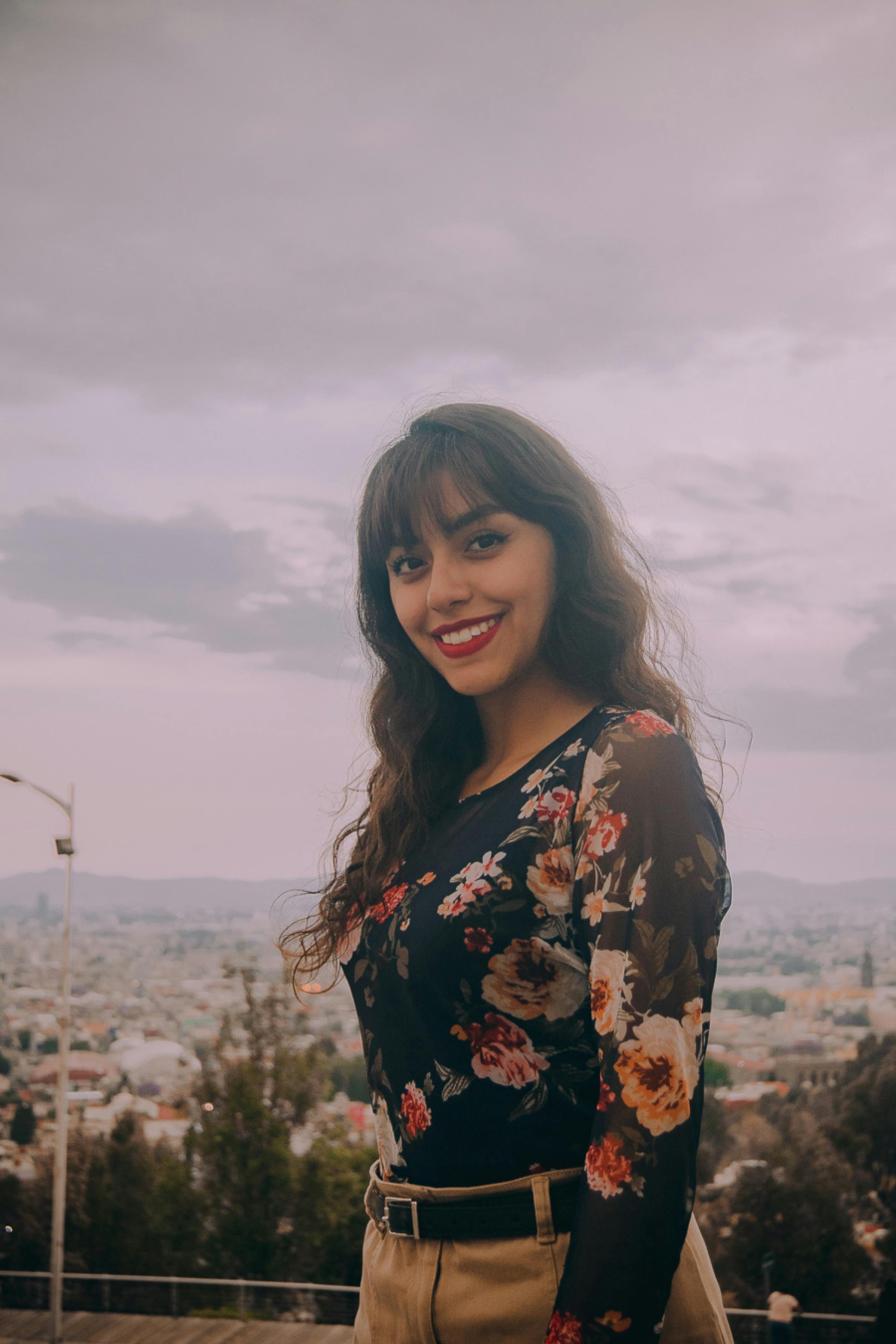 Smiling woman in a floral top with a cityscape view in the background, captured outdoors.