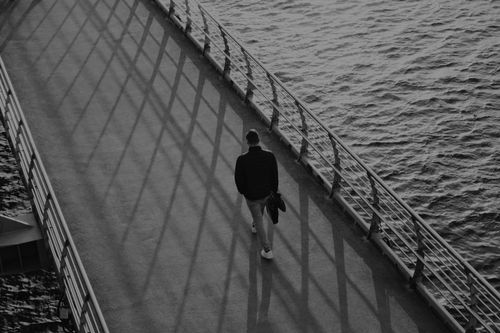 Free A man walks alone on a boardwalk by the water, casting long shadows. Stock Photo