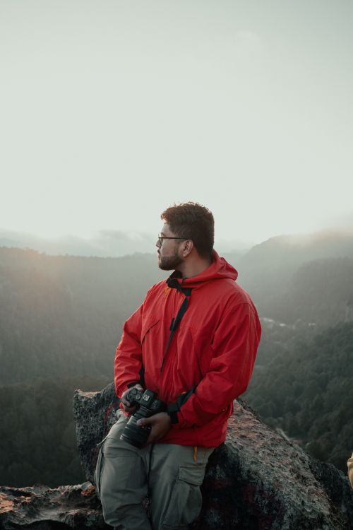 Free Photographer in red jacket sitting on rocky cliff with scenic mountain view. Stock Photo