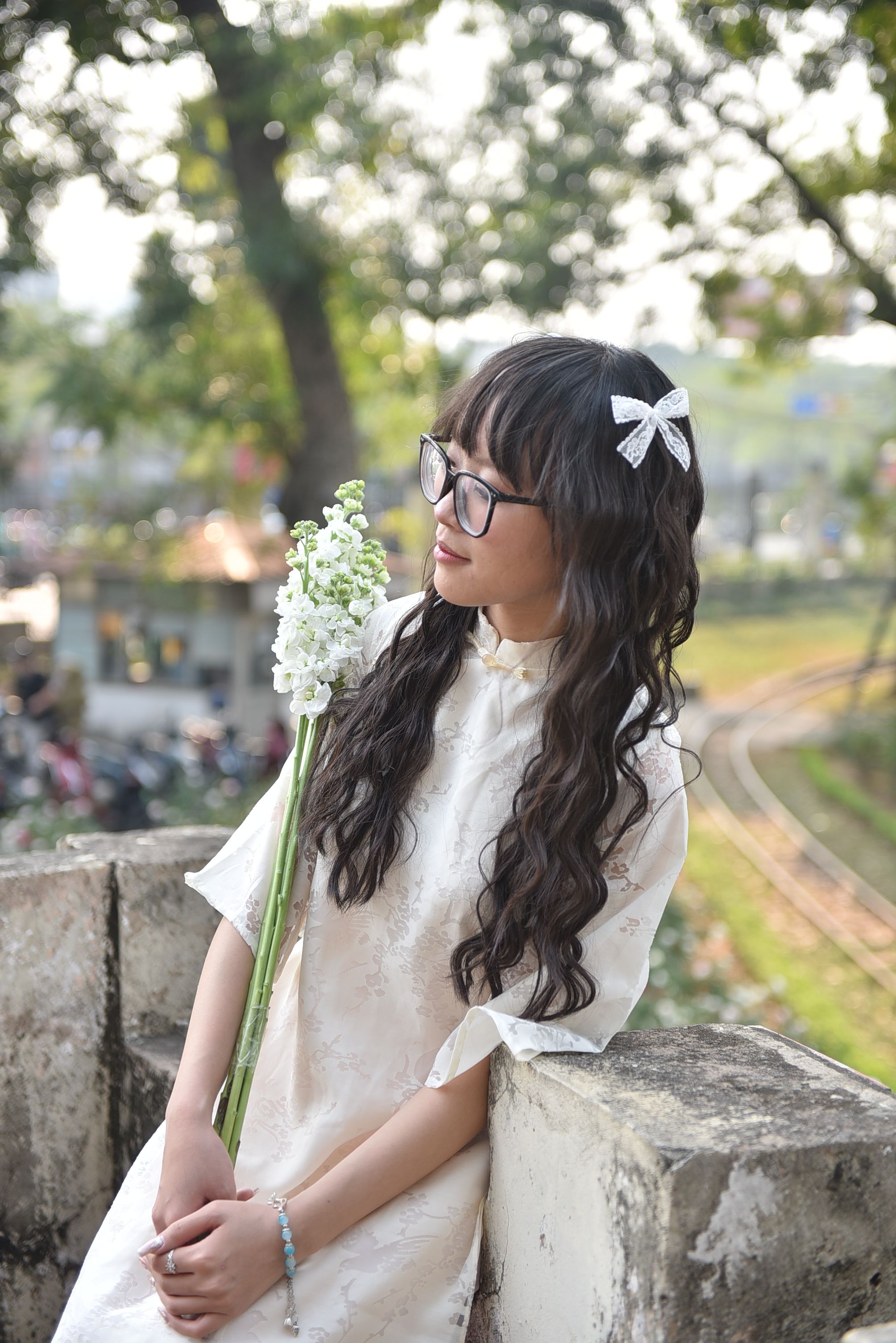 Young woman with glasses holding flowers, wearing a white dress, outdoors.