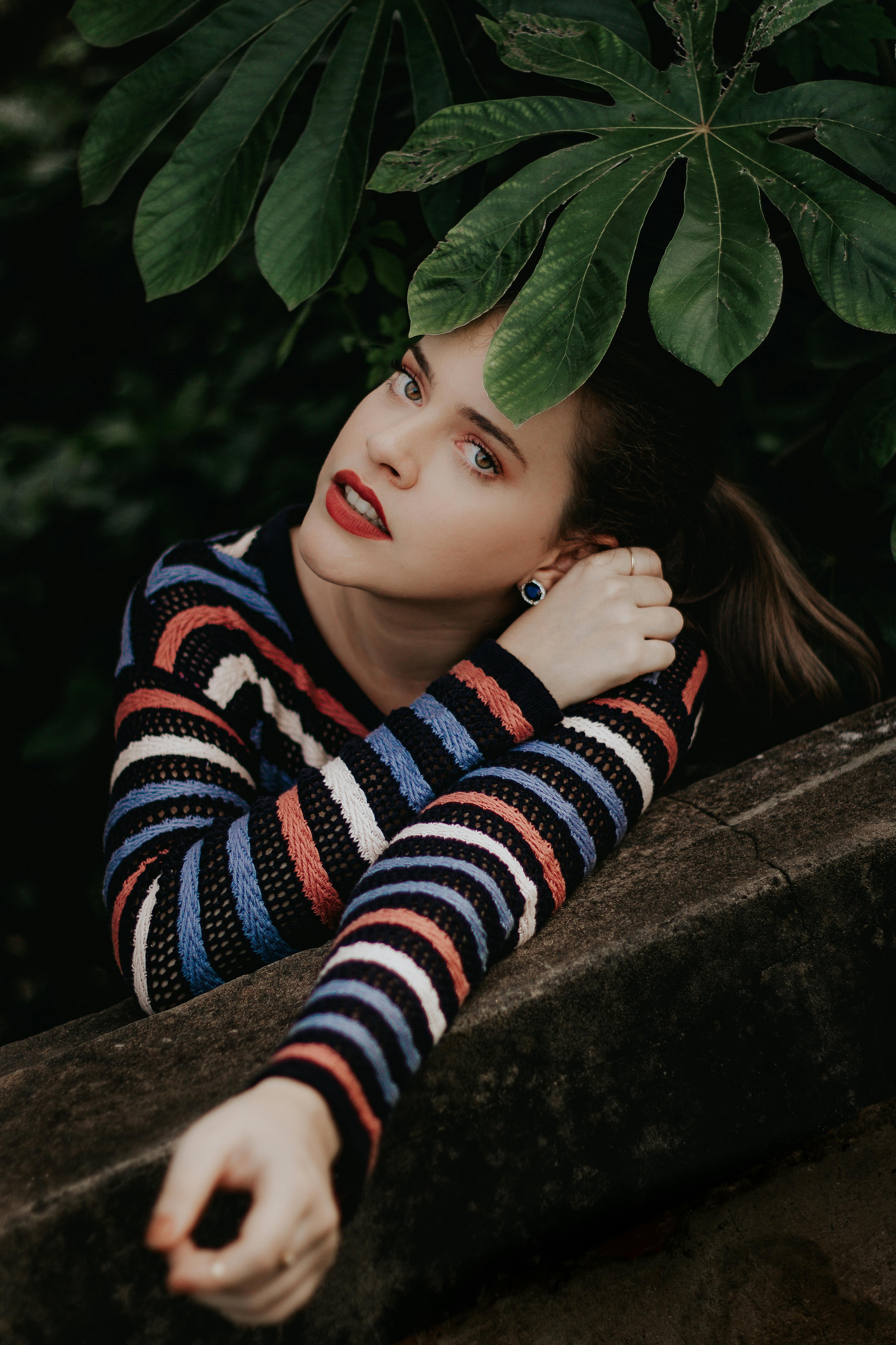 一位身穿活力条纹毛衣的女子，倚靠在石墙上，四周绿叶环绕