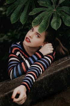 一位身穿活力条纹毛衣的女子，倚靠在石墙上，四周绿叶环绕