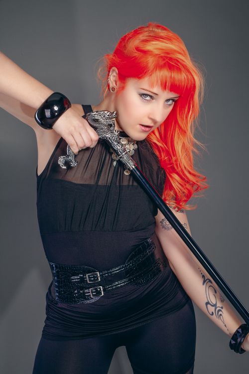 Free Confident woman with bright red hair and a stylish black outfit holding a decorative sword. Stock Photo