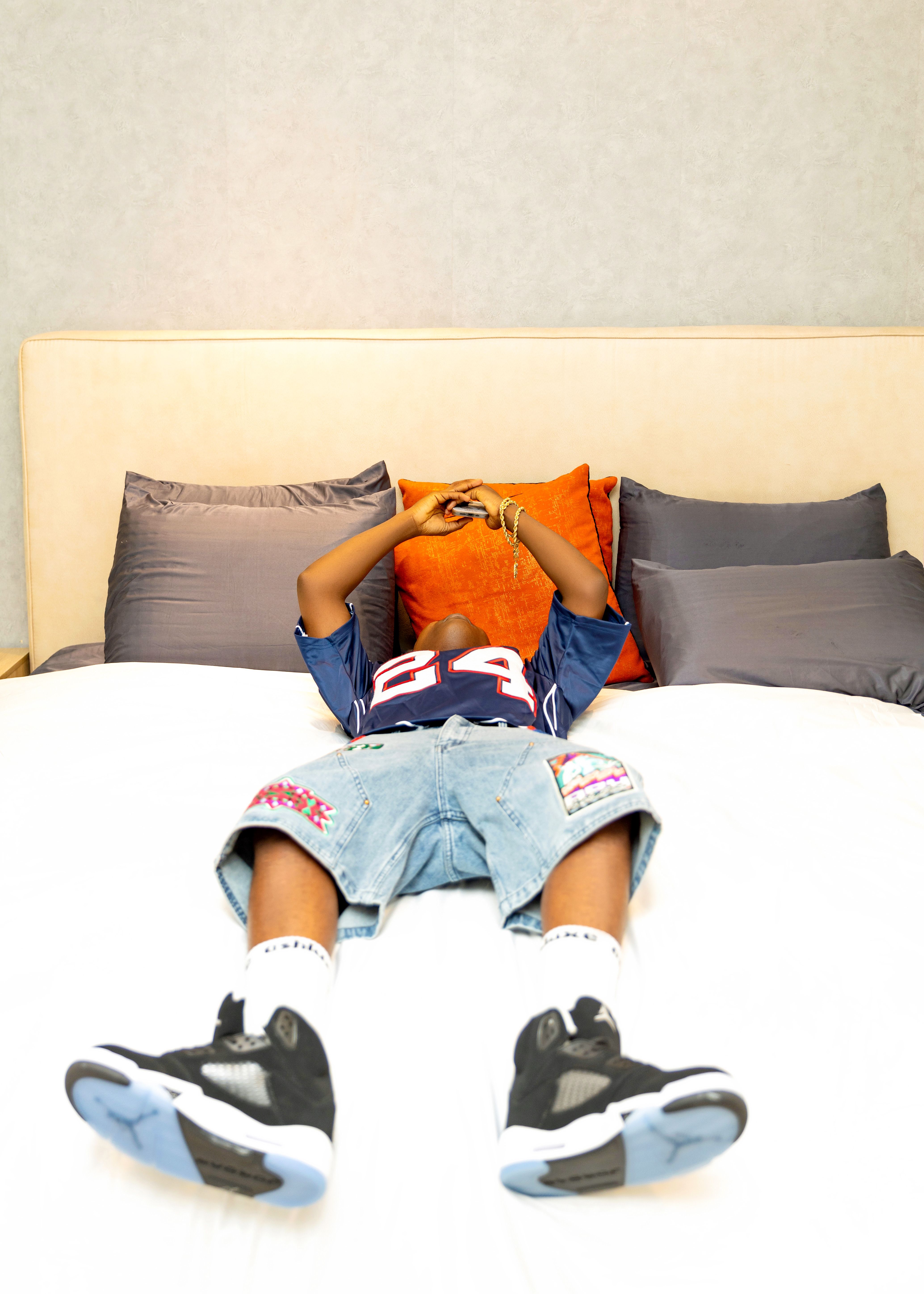 Teenage boy wearing a jersey and sneakers, relaxing on a bed indoors.