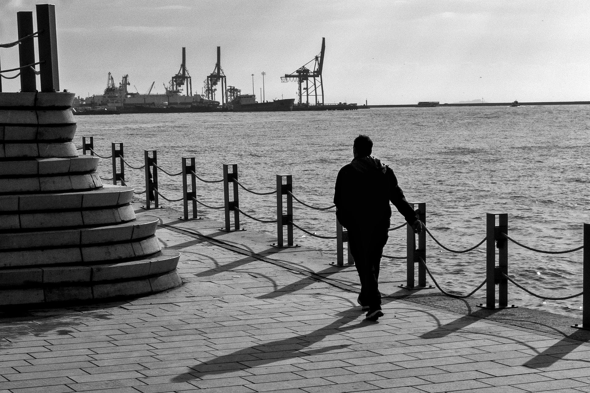 A silhouette of a person walking along a waterfront with industrial cranes in the background, creating a moody scene.