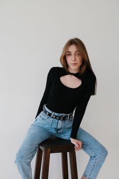 Stylish young woman in black top and jeans sitting on stool against plain background.