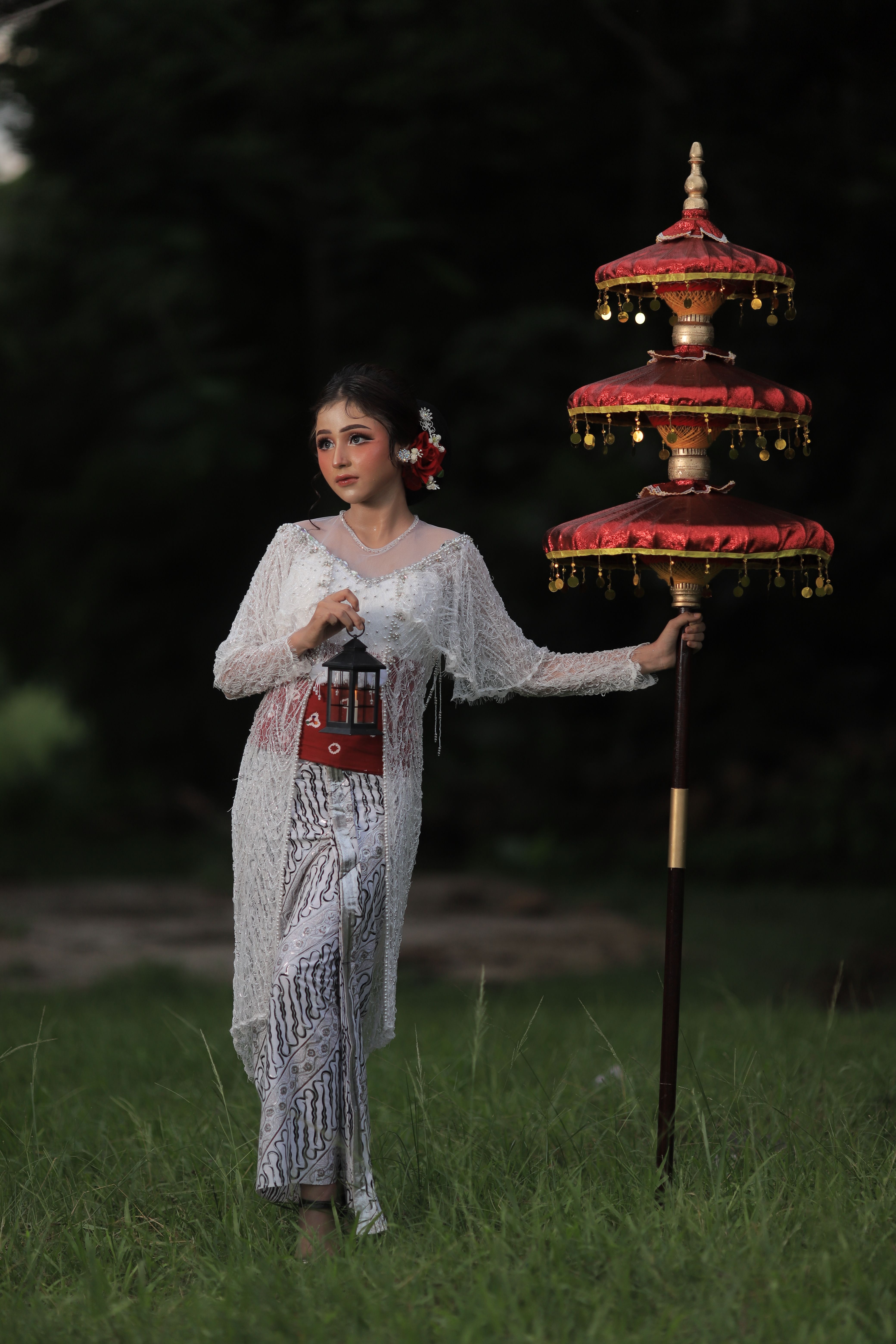 A woman in traditional clothing stands in a serene outdoor setting, holding a decorative fan.