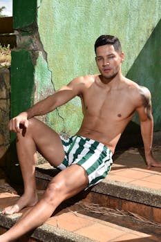 Young man in swim shorts sitting casually on outdoor steps under summer sunlight.