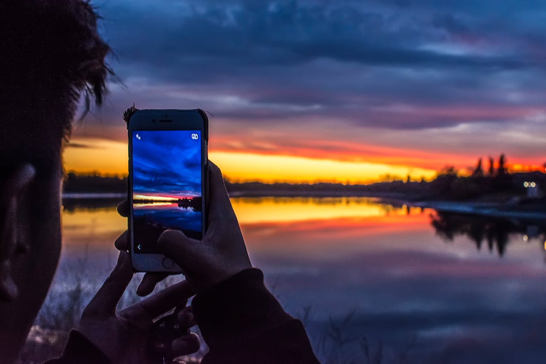 無料 ゴールデンアワーの下で湖の写真を撮るスマートフォンを持っている人 写真素材