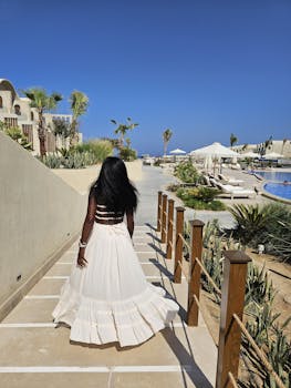 Back view of a woman in a white dress walking by a luxurious resort poolside in sunny weather.