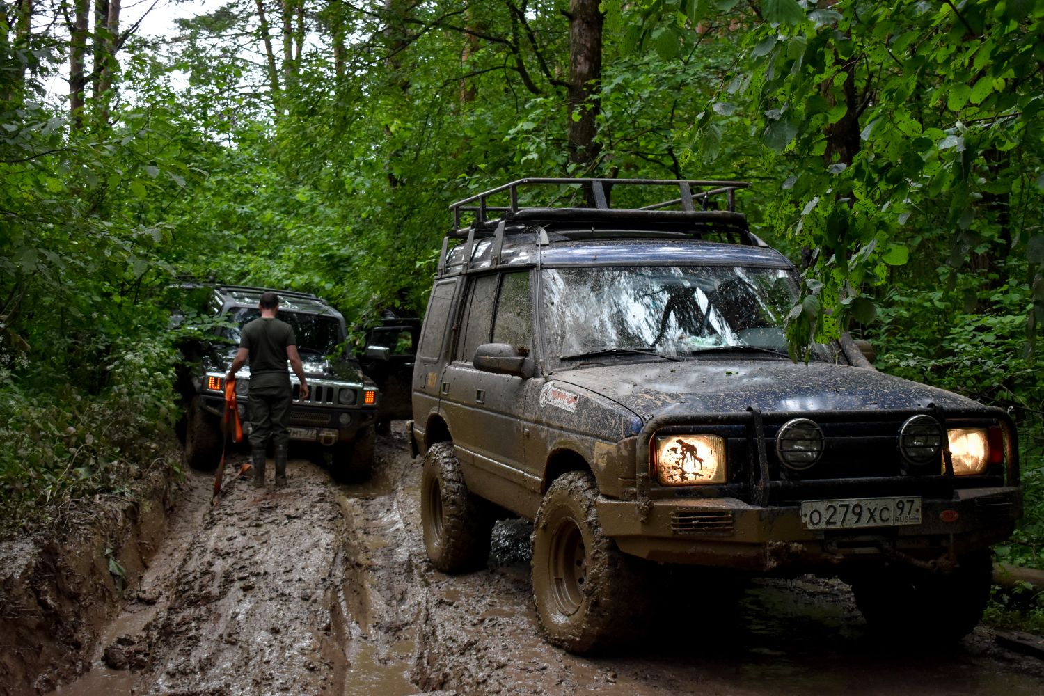 Two mud-covered off-road SUVs on a deeply rutted forest trail with a recovery operator preparing a strap between them