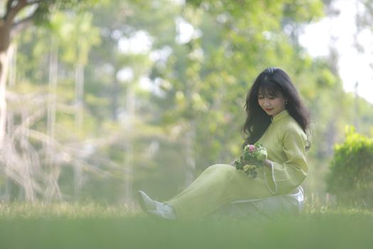 A serene woman in traditional attire sits on grass holding flowers in a lush, sunlit park.