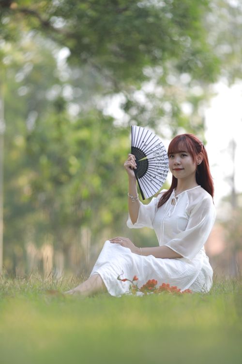 Free A serene portrait of a young woman in a white dress holding a fan, seated on grass. Stock Photo