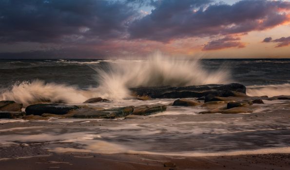 Powerful ocean wave splashing over rocks during sunset, with dramatic clouds.