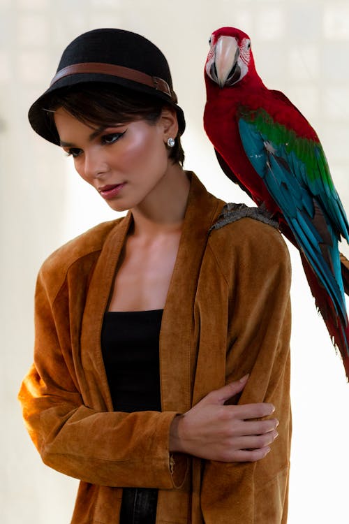 Young serious female with glowing makeup standing with exotic bird on shoulder and looking down