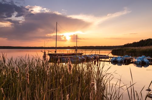 Free Tranquil lake scene with boats docked, set against a vibrant sunset and surrounded by reeds. Stock Photo