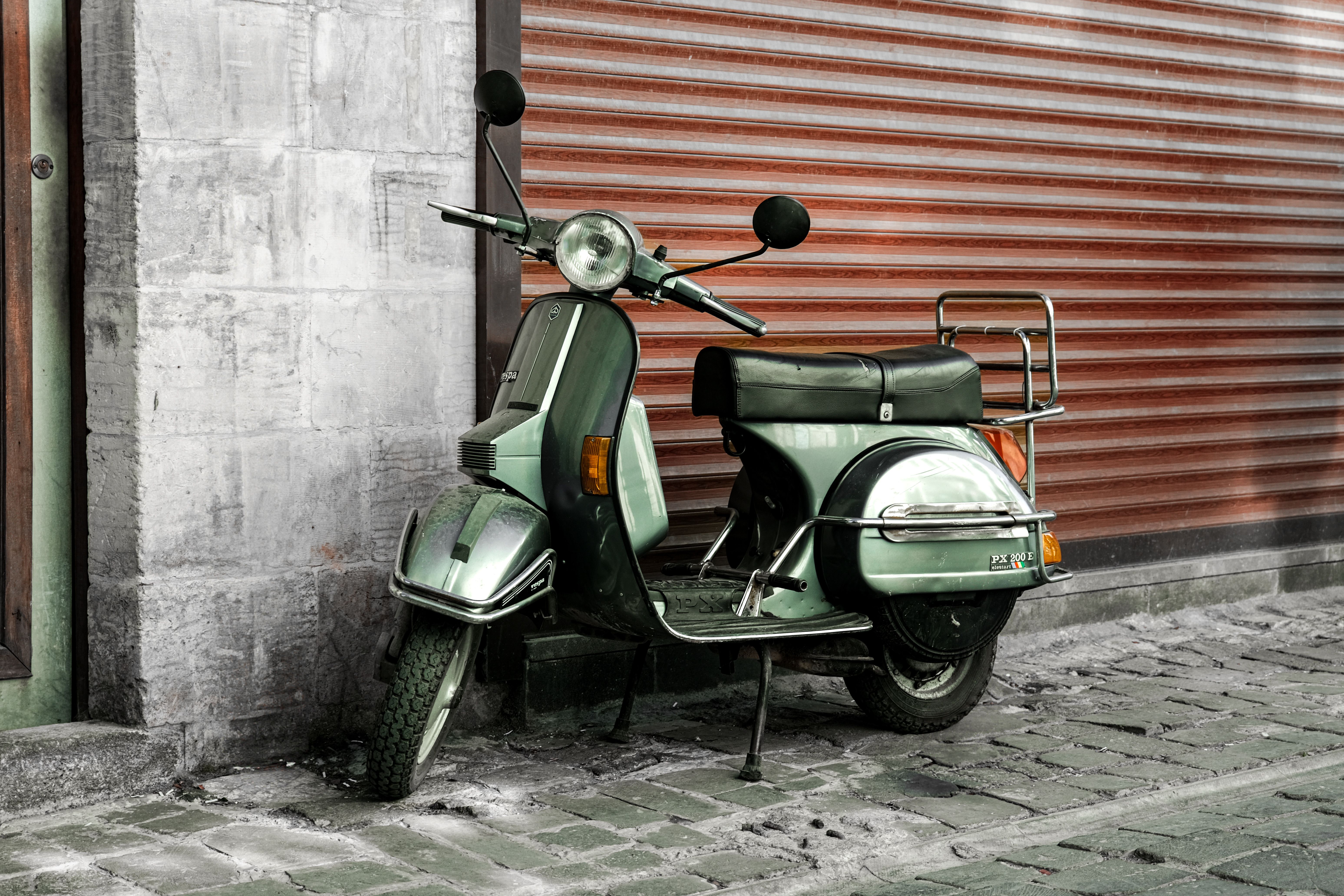 Classic green scooter parked against a metal shutter in a cobblestone alleyway.