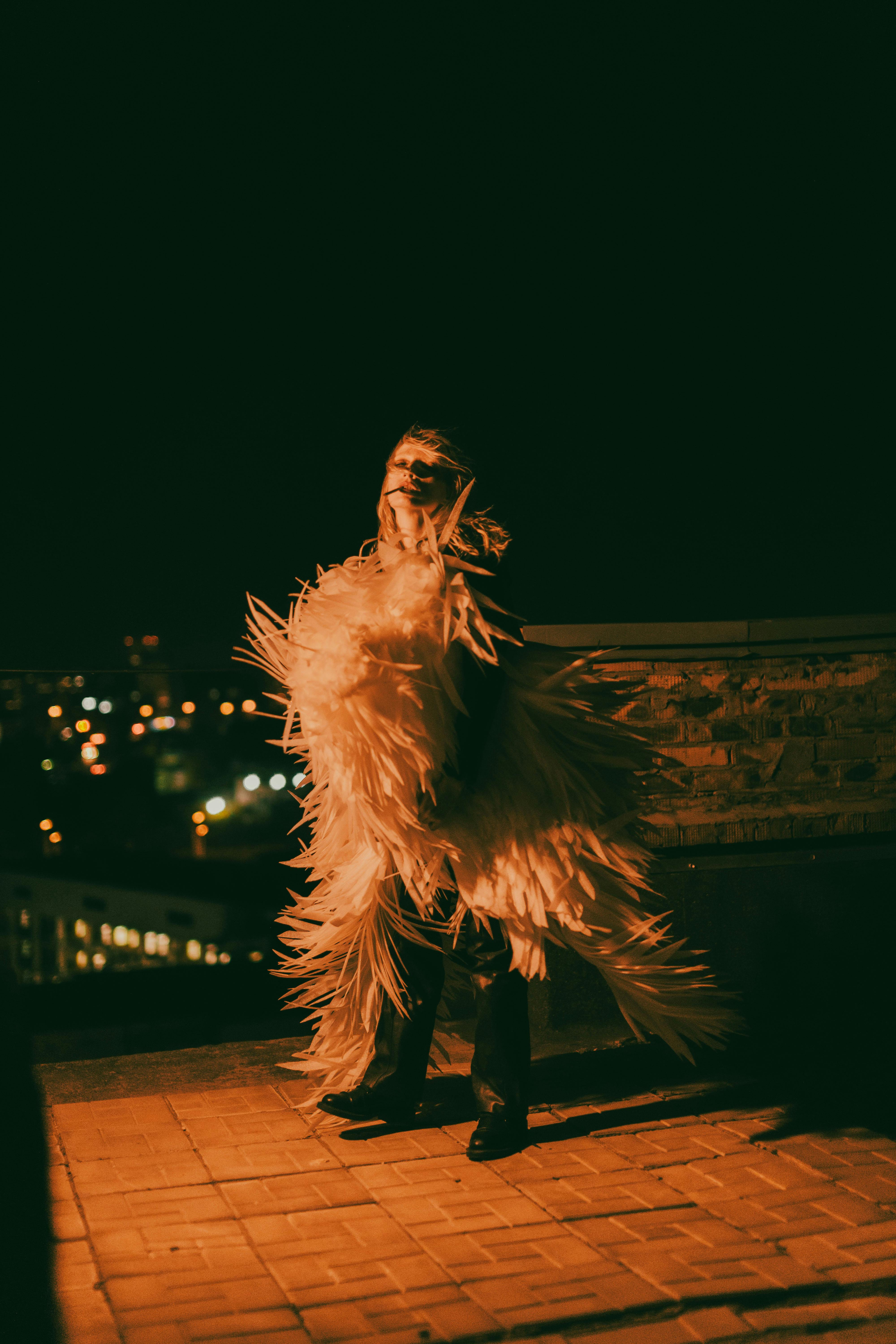 Kostenlos Ein dramatisches Nachtporträt einer Person in einem extravaganten Federkostüm vor einer Stadtlandschaft. Stock-Foto