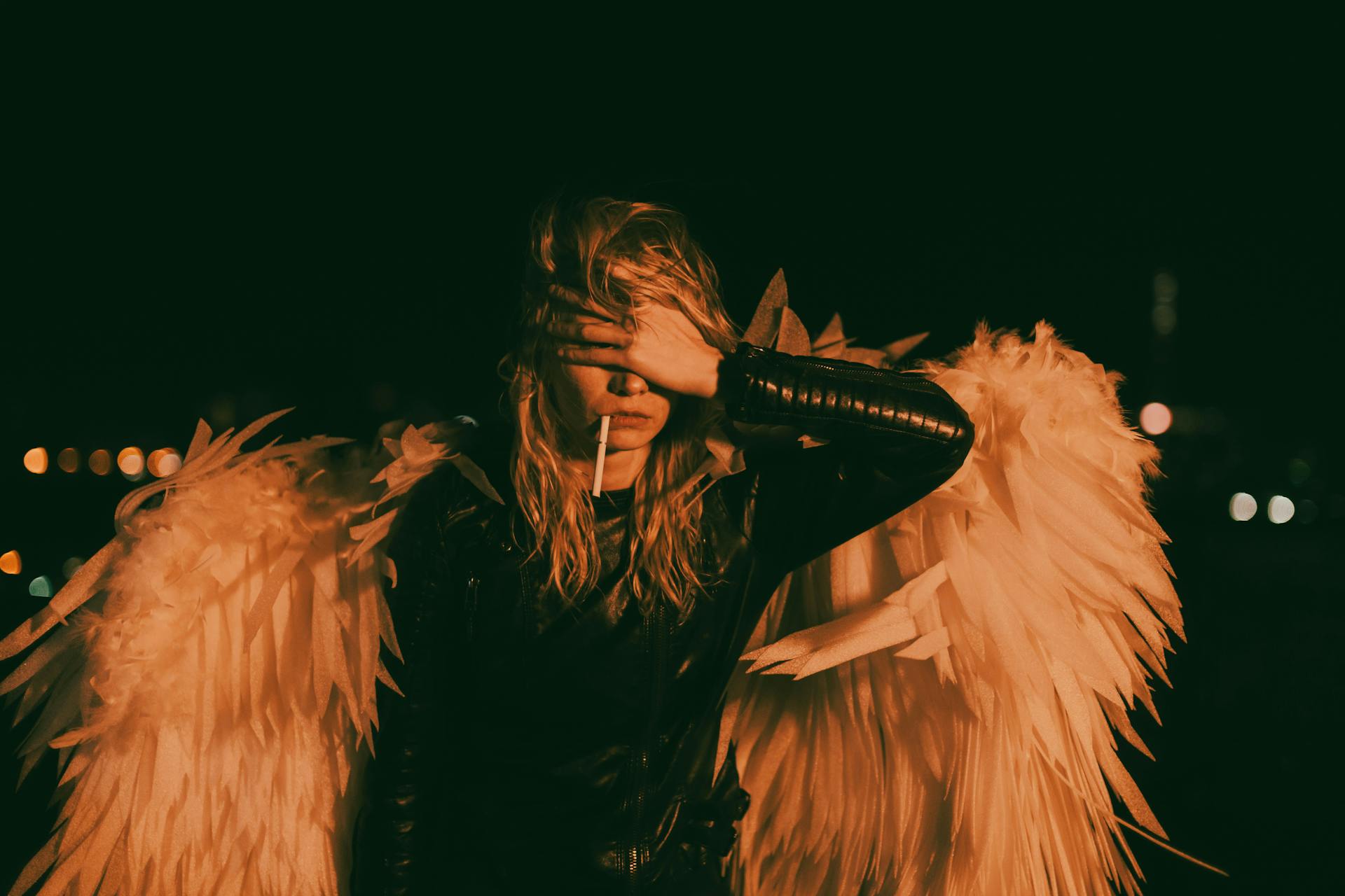 A person dressed as an angel with wings and blonde hair covers their face in a dark urban setting.