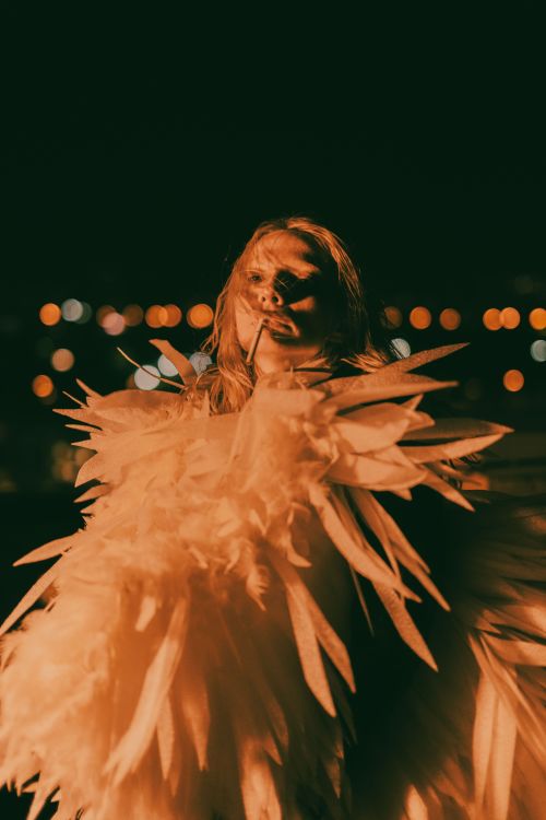 Free A dreamy portrait of a person adorned in feathers with glowing city lights at night. Perfect for fashion and artistic themes. Stock Photo