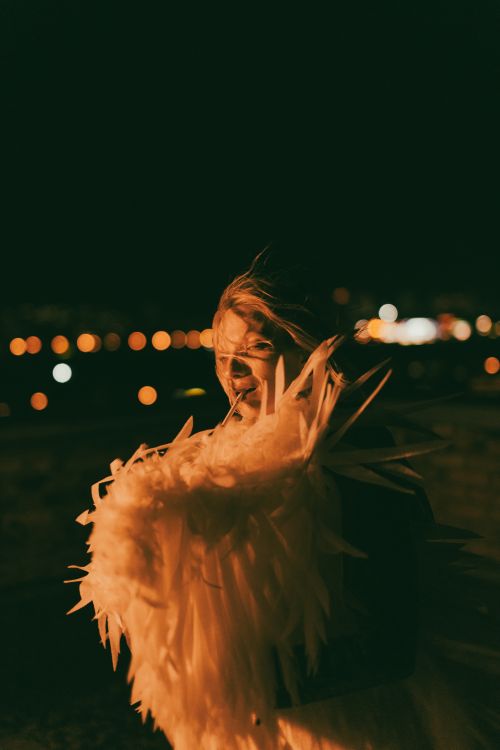 Free Captivating nighttime portrait of a person with dramatic feathered attire and city lights in background. Stock Photo