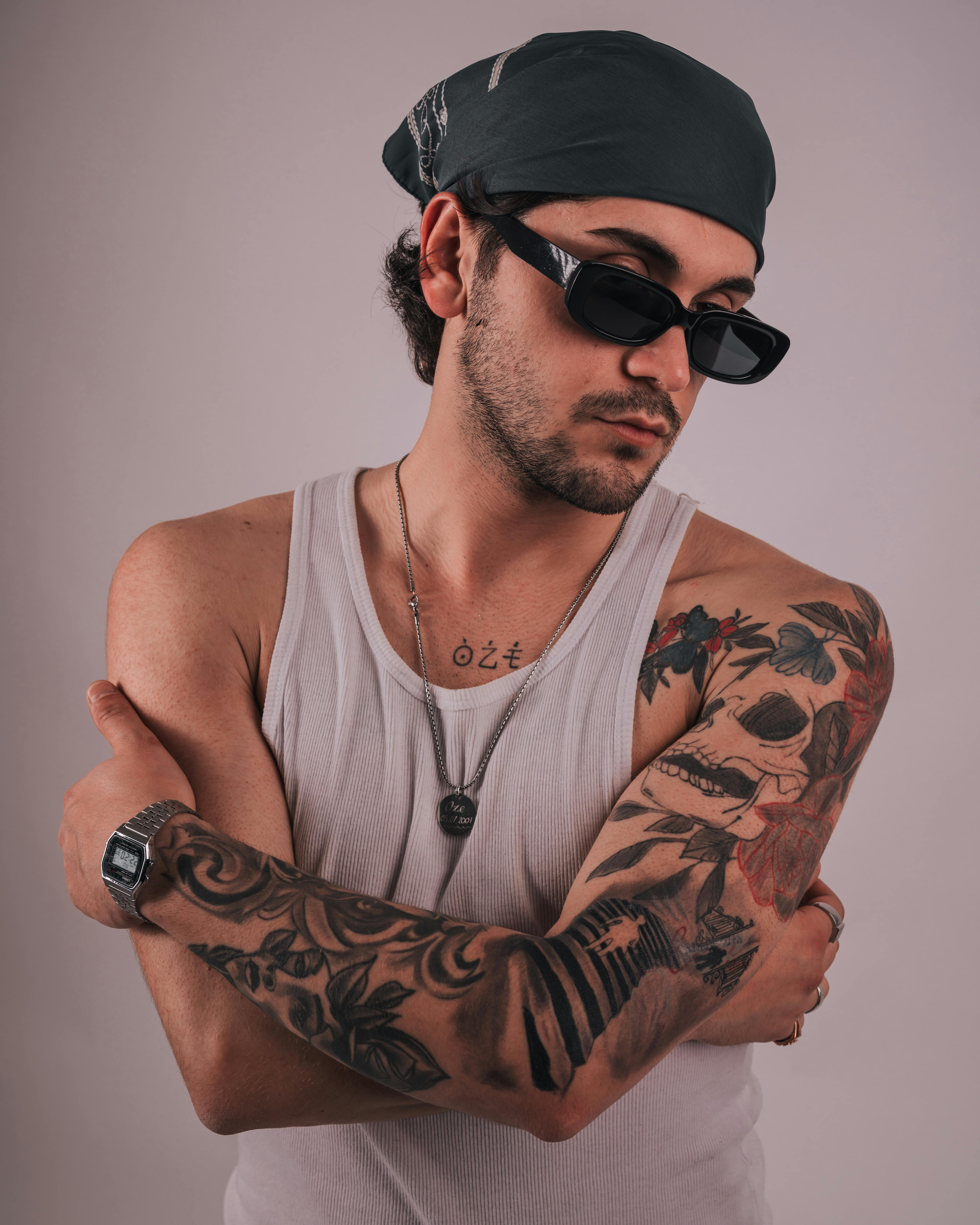 Young adult male with tattoos wearing sunglasses and bandana posing with arms crossed.
