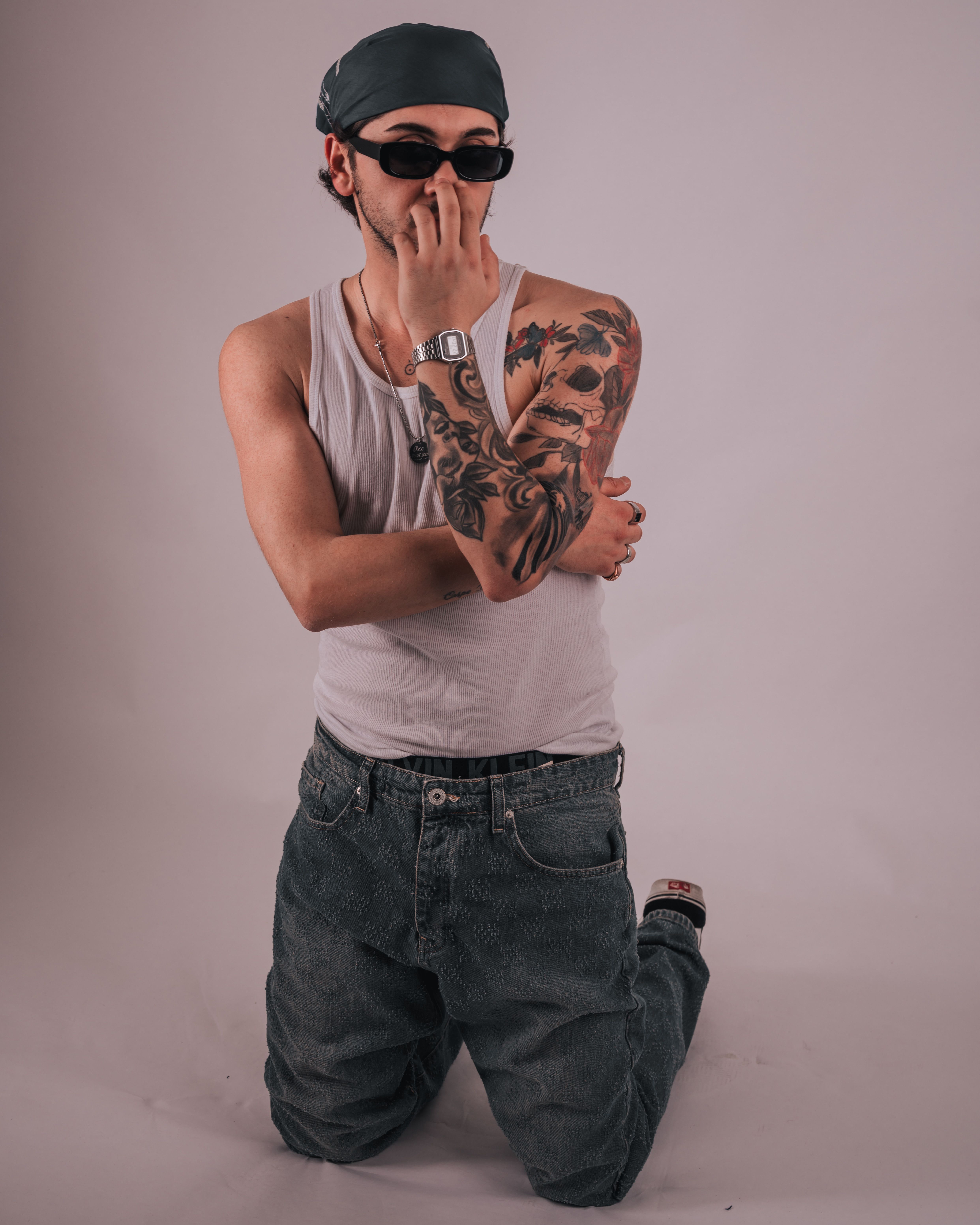 Trendy man kneeling indoors, showcasing tattoos and urban fashion style.