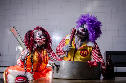 Creepy clowns in makeup and costumes with horror props in a kitchen scene.