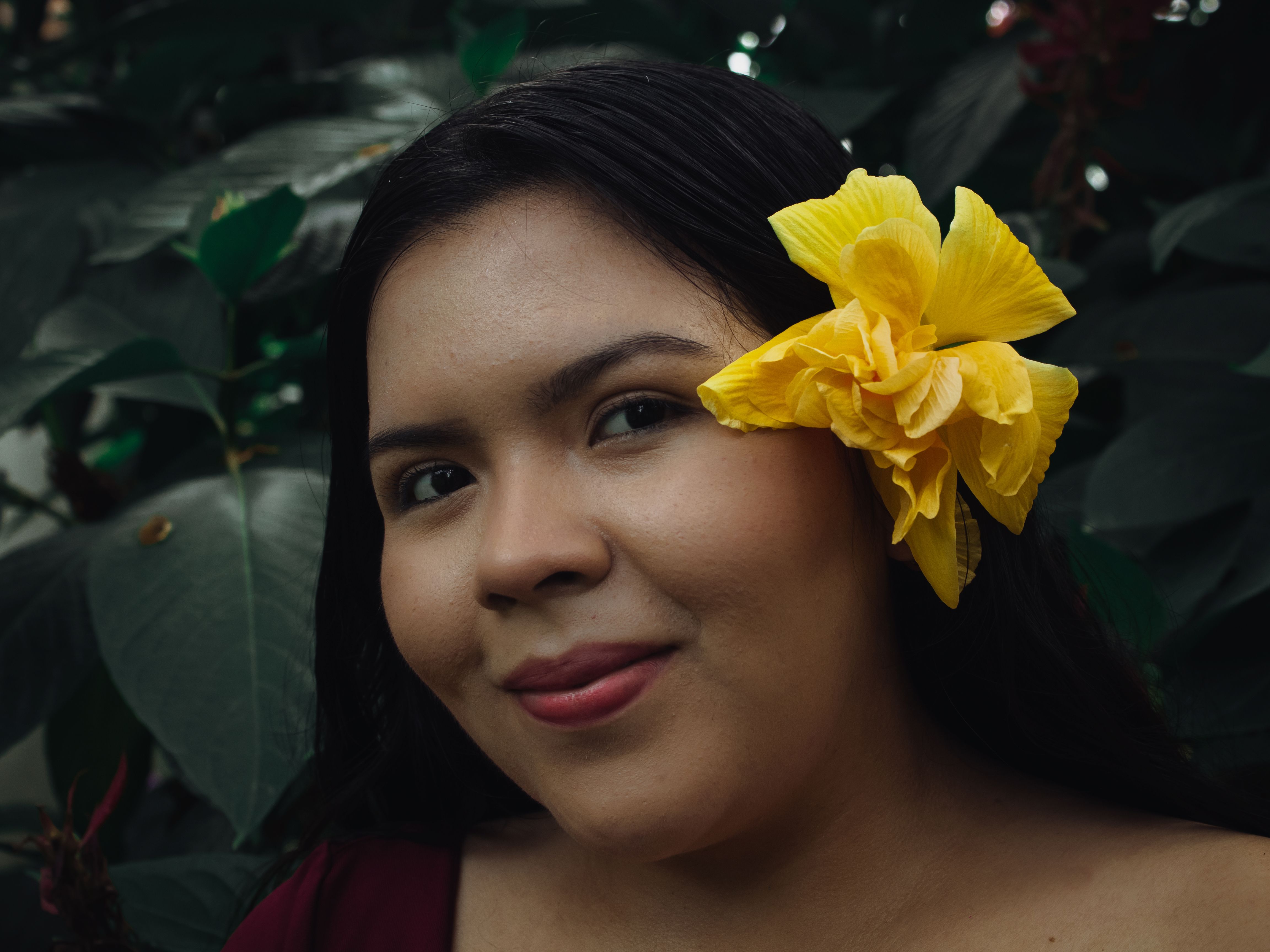 Close-up portrait of a woman with a yellow flower in her hair surrounded by green leaves.